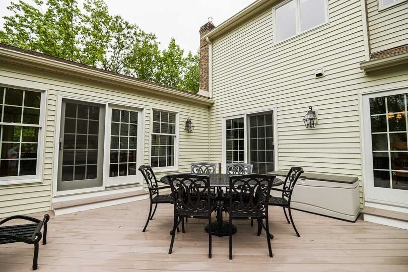 A dining table and chairs sit on a light-colored wooden deck attached to the back of a beige, two-story vinyl house.