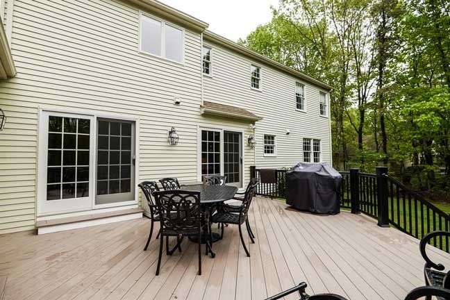 A light-colored house exterior with a wooden deck featuring a dining table, chairs, and a covered grill.