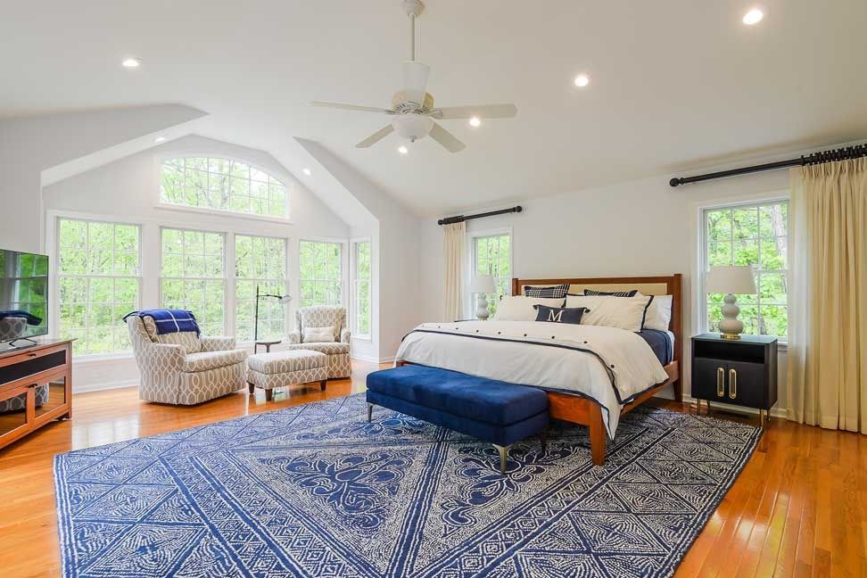 A bright, spacious bedroom with hardwood floors, a large blue patterned rug, a bed with a blue bench, and a seating area.