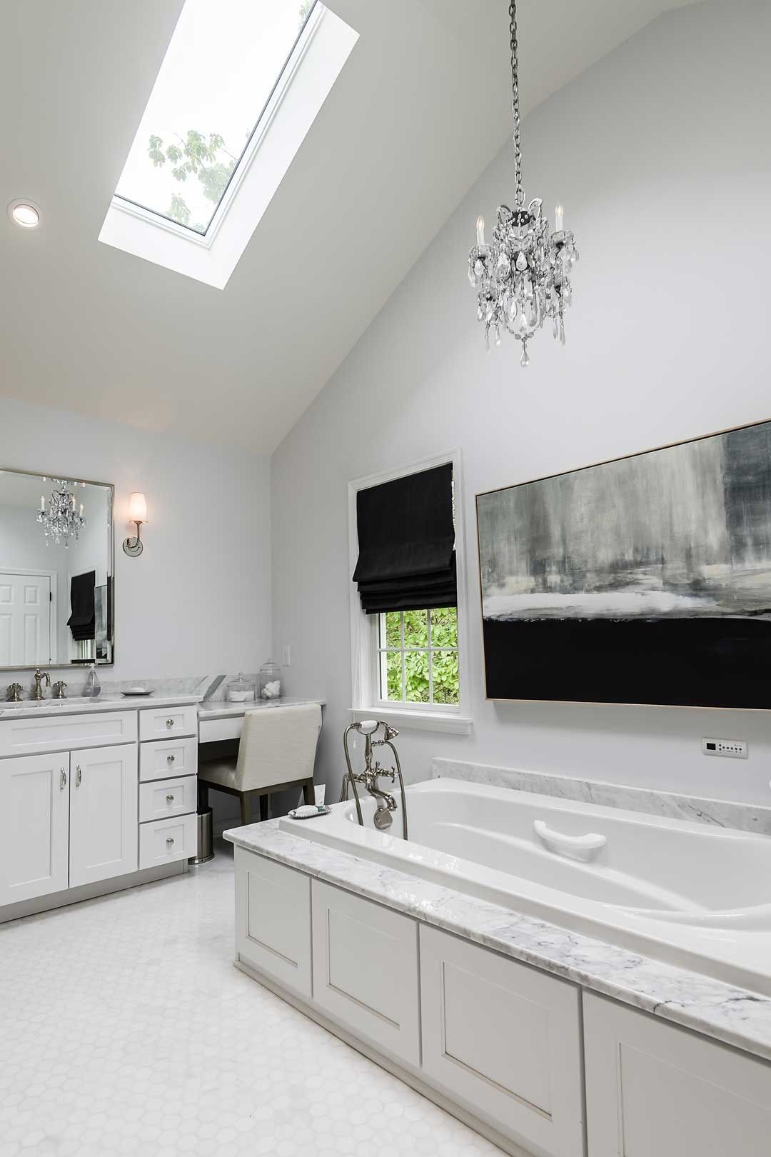 A bright, modern bathroom featuring a white vanity, built-in bathtub, a crystal chandelier, and a skylight.