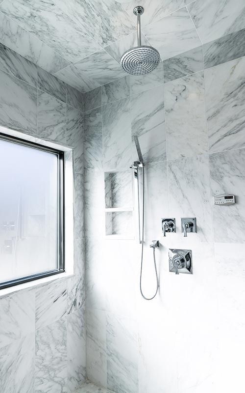 A modern shower stall featuring white marble wall and ceiling tiles, a rainfall showerhead, and a hand-held sprayer.