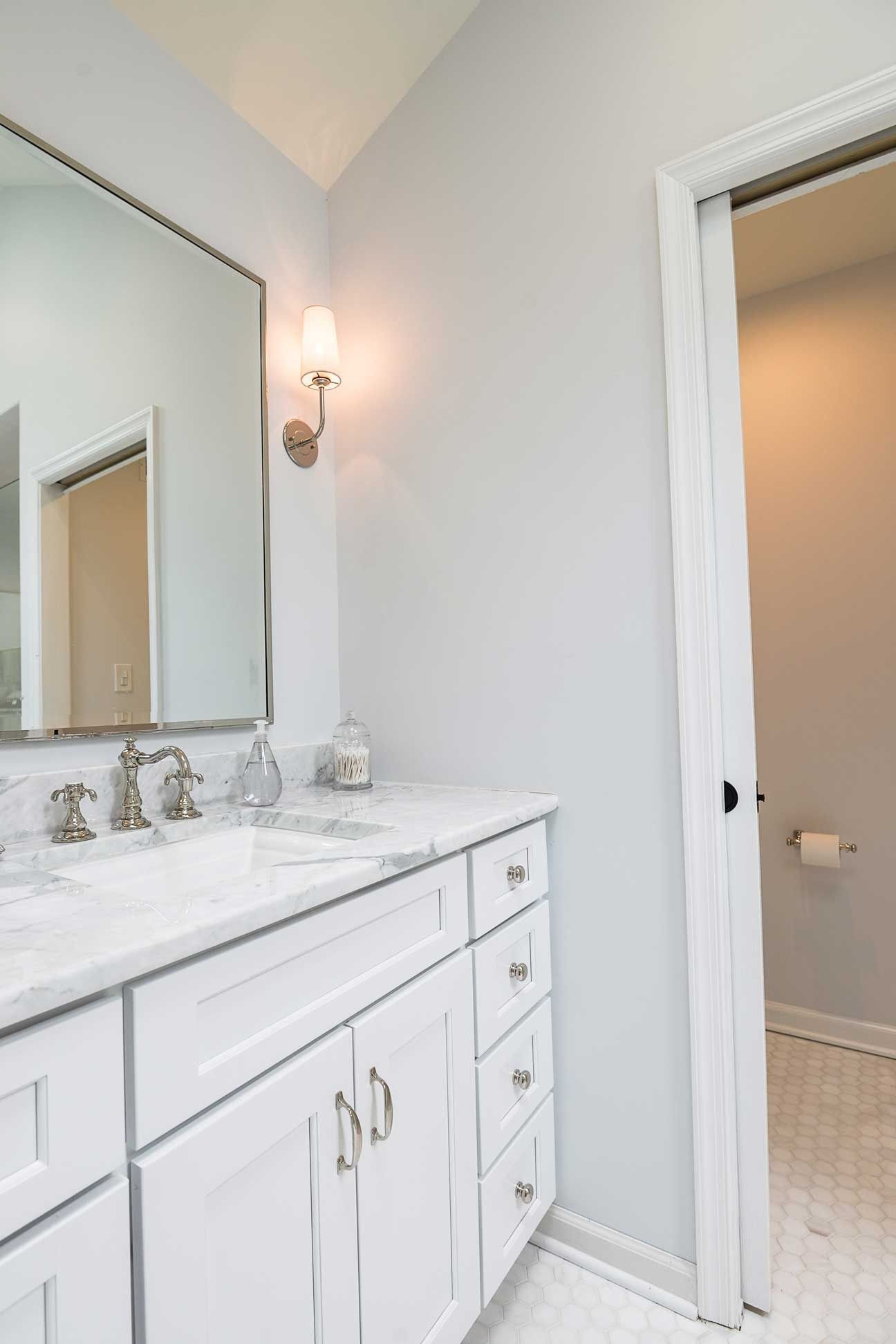 A bright, modern bathroom featuring a white vanity with marble countertop, a large mirror, and a door to an adjacent room.