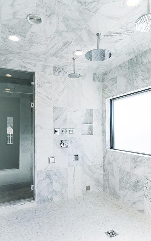 A luxurious shower with light gray marble walls, multiple showerheads, a glass door, and a window.