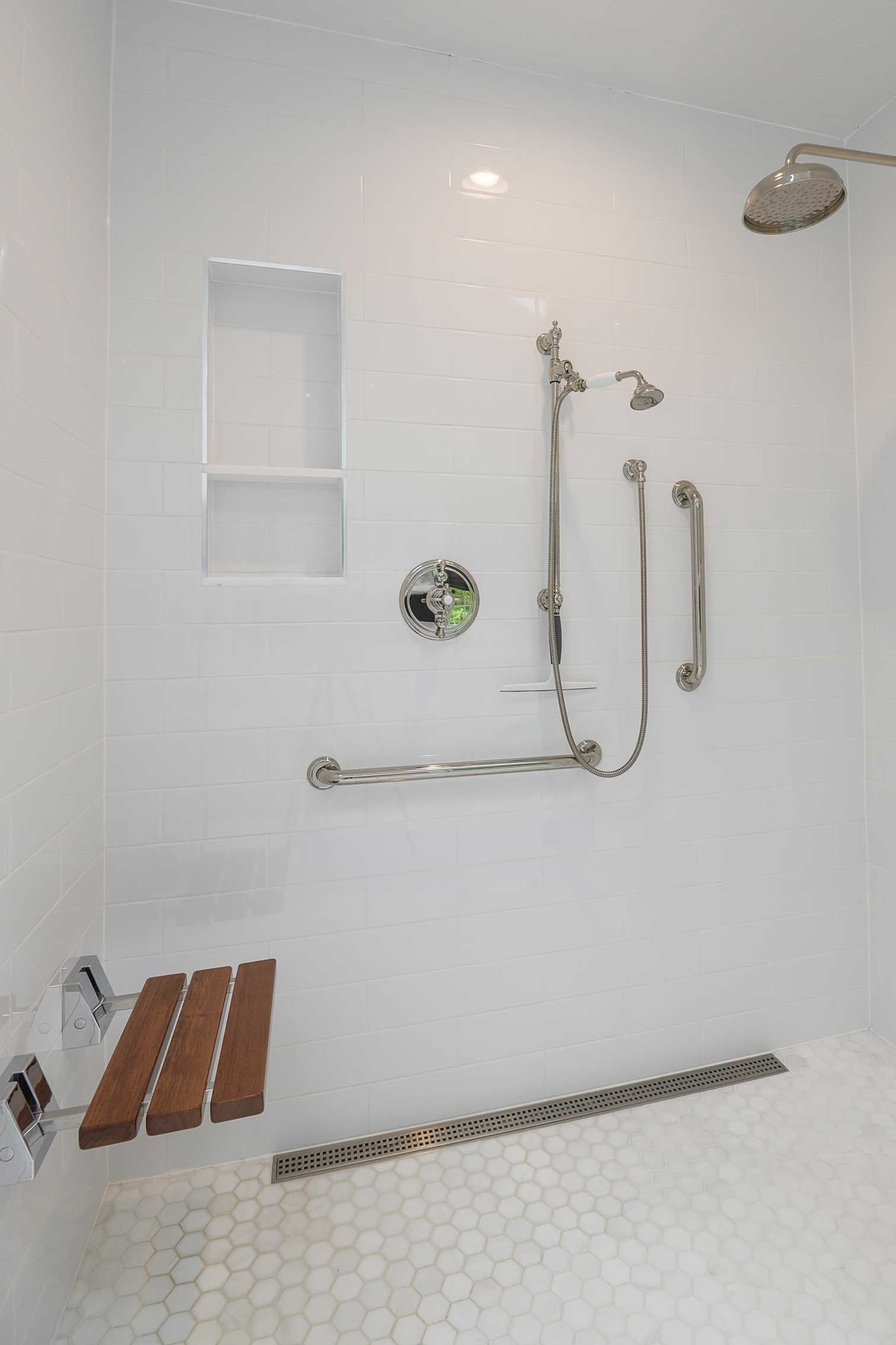 An accessible walk-in shower with a white tiled interior, wooden fold-down seat, safety grab bars, and a linear drain.