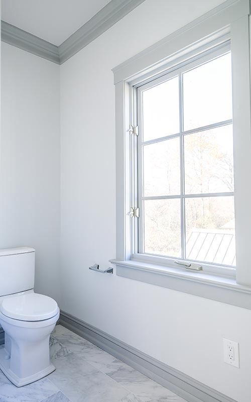 A minimalist bathroom featuring white walls, gray trim and molding, marble-style flooring, and a white toilet by a window.