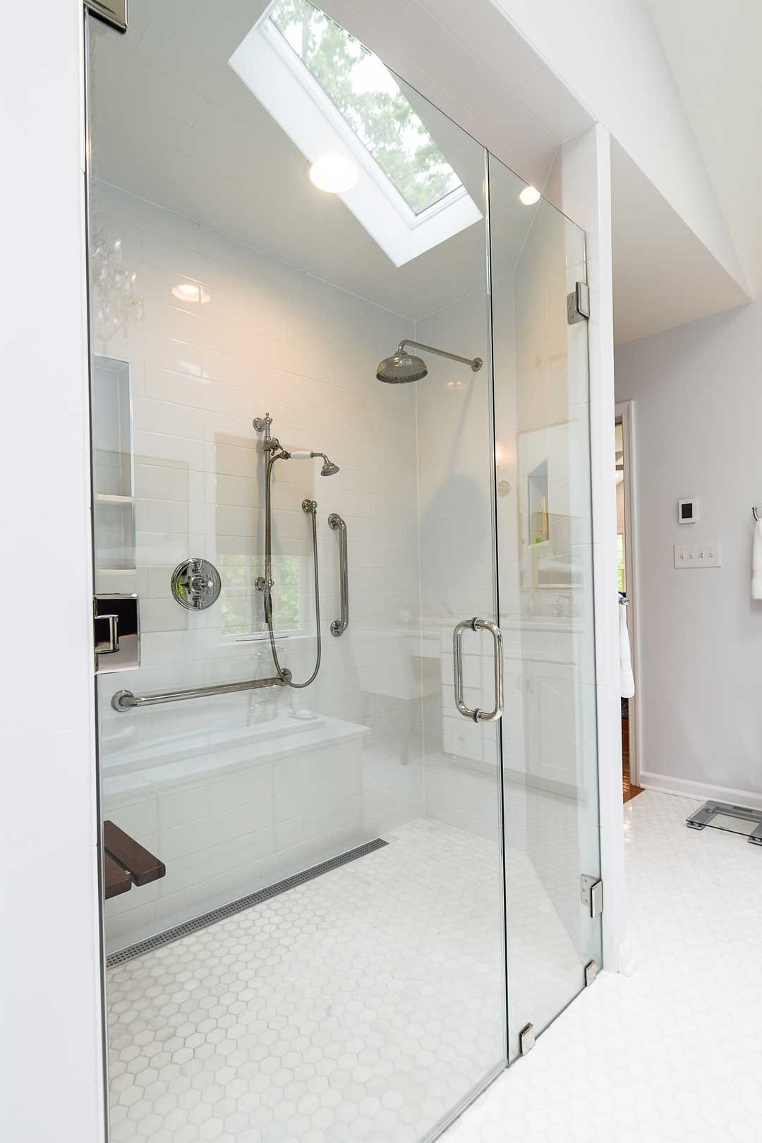 A modern walk-in shower with a glass door, white tiled floor, grab bars, a built-in bench, and a skylight above.