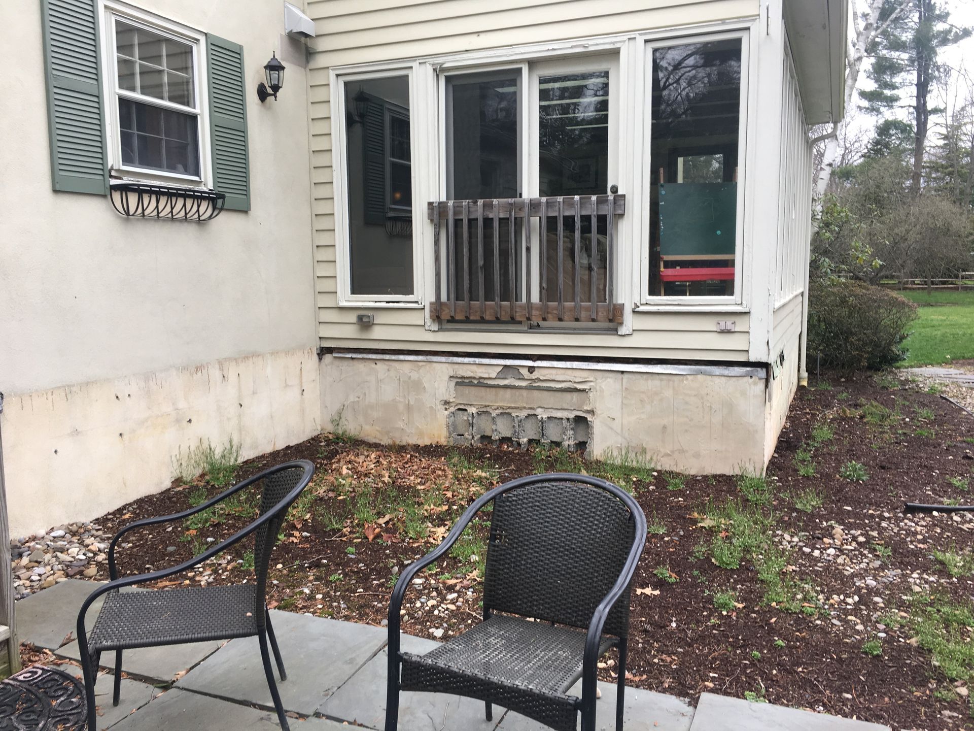Two black chairs on a stone patio in front of a house with a beige porch, sliding glass doors, and a concrete foundation.