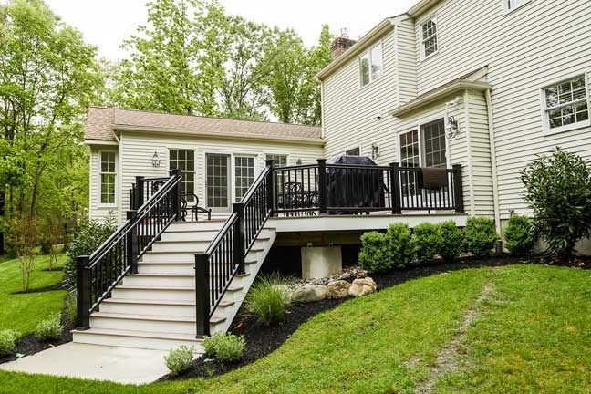 Light-colored suburban house with a large composite deck and staircase leading to a manicured lawn and landscaping.