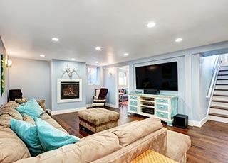A cozy living room with a tan sectional, a light-blue fireplace wall, a flat-screen TV on a console, and basement stairs.