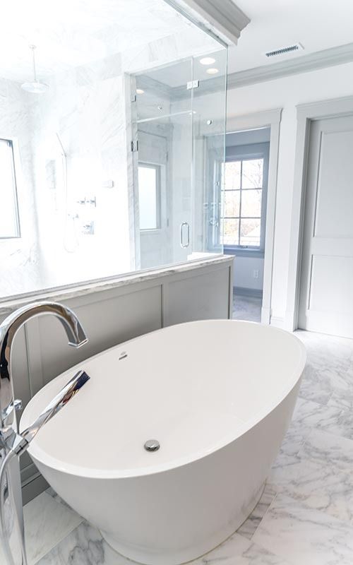 A freestanding oval bathtub sits in a modern, bright bathroom with marble floors and a glass-enclosed shower.