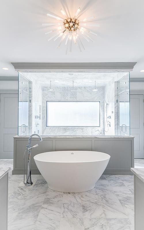 A freestanding white soaking tub sits in front of a marble-tiled wall with a central window and overhead starburst light.