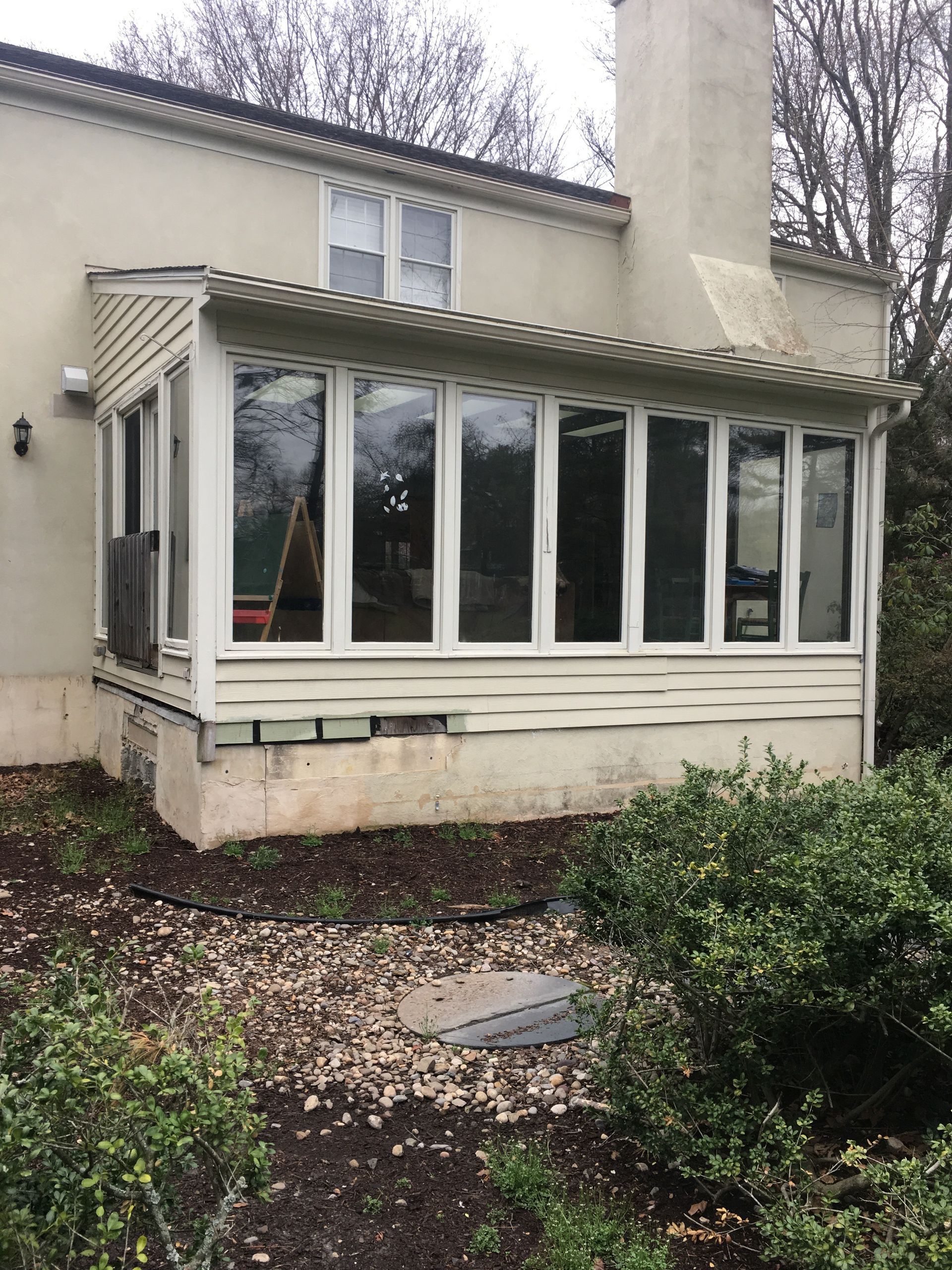 A sunroom addition with white-framed windows, light siding, and an exposed concrete foundation attached to a stucco house.