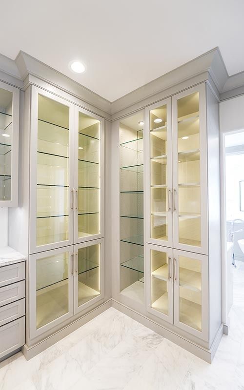 Light-gray corner cabinetry with glass-fronted doors, interior lighting, and shelves over white marble floors.