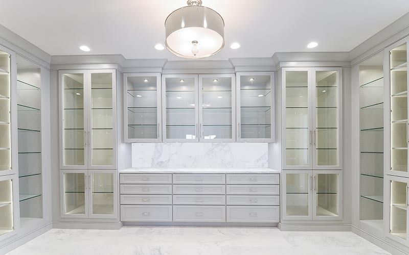 A luxurious walk-in closet featuring light gray cabinetry with glass-front display shelves, drawers, and a marble top.