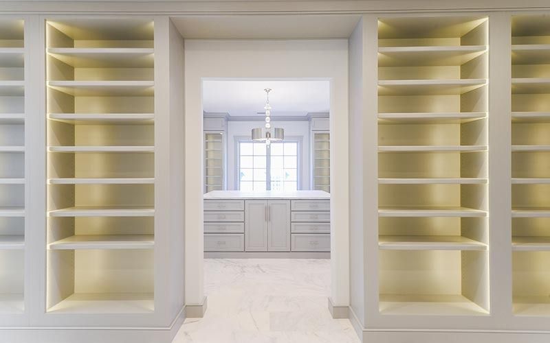 A symmetrical view of a hallway lined with backlit, built-in shelving, leading to a bright, modern dressing area.