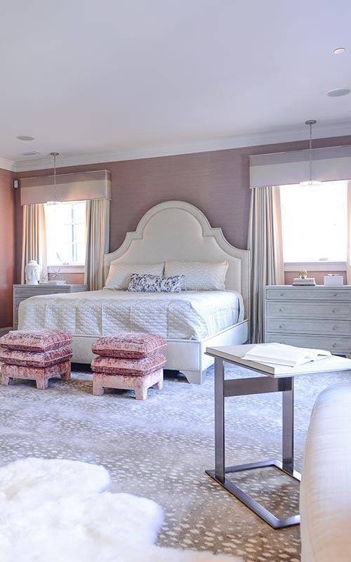 A bedroom featuring a large white arched headboard, two matching dressers, mauve accent walls, and two floral footstools.