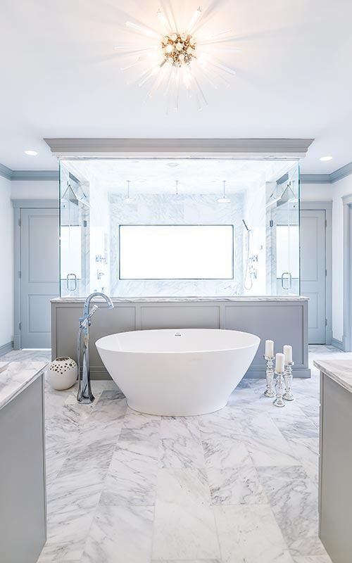 A luxurious master bathroom featuring a white freestanding tub, marble floors, grey cabinetry, and a large central mirror.