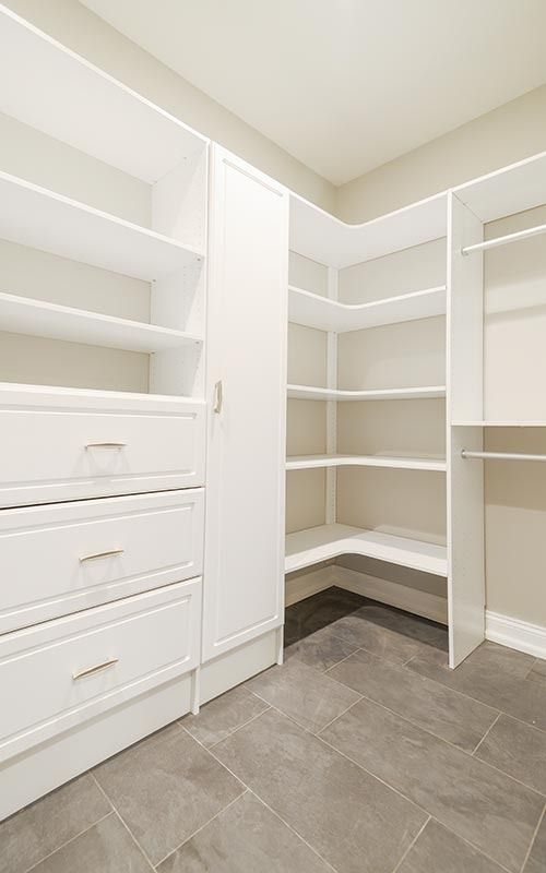 A walk-in closet featuring white wooden shelving, drawers, and hanging rods over a gray tiled floor in a neutral room.