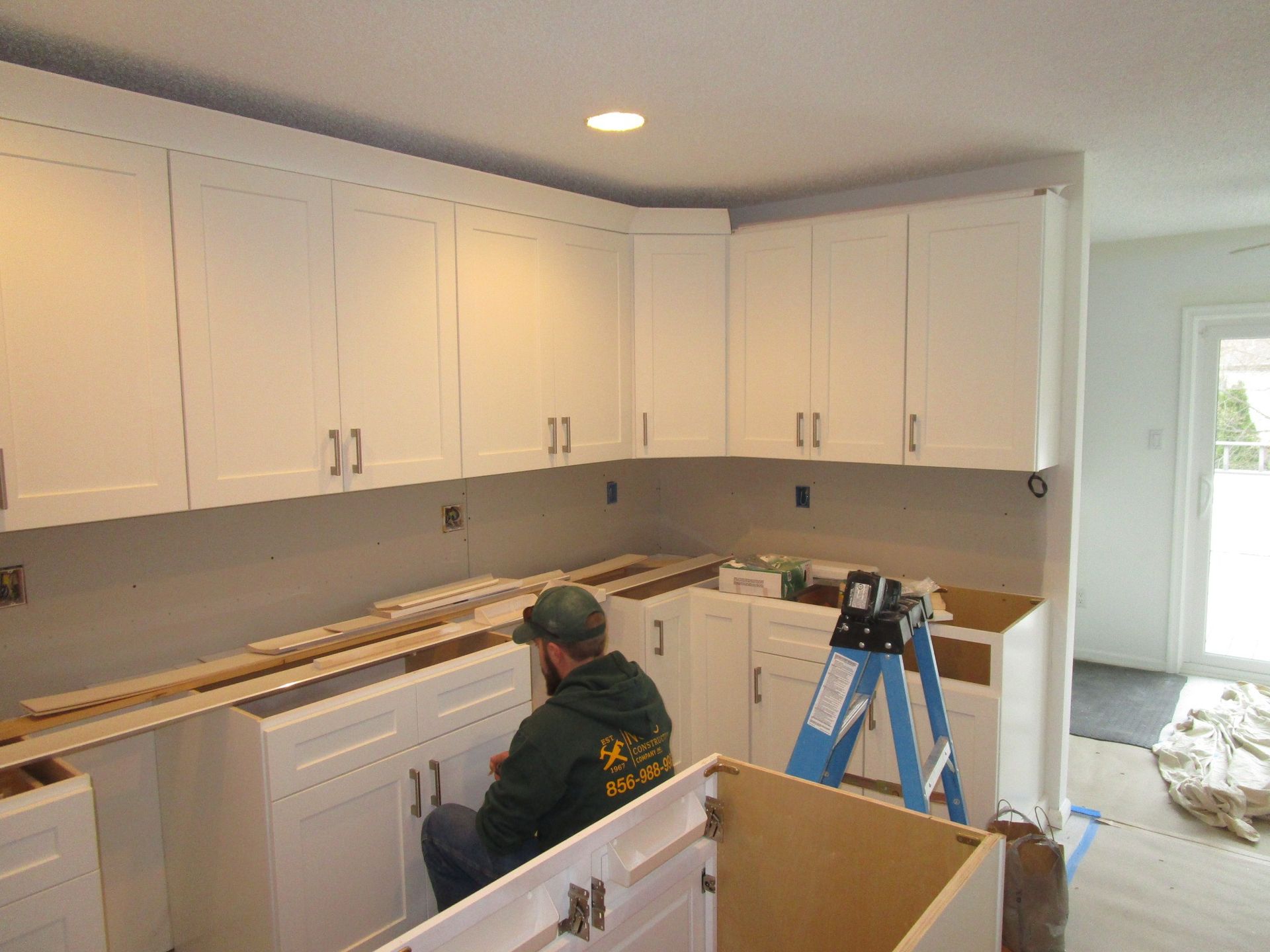 A person in a dark jacket installs white kitchen cabinets and countertops in a home under renovation.