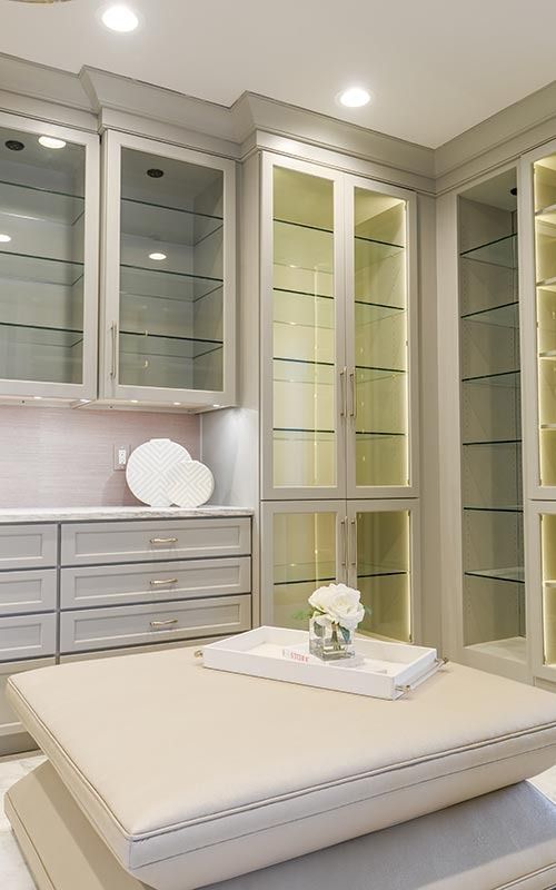 A modern, light gray walk-in closet with illuminated glass-front cabinets, stacked drawers, and a central ottoman.