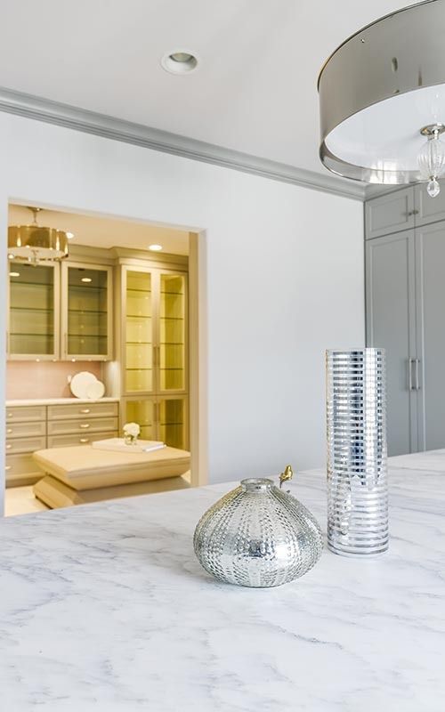 A marble kitchen island with a silver vase and decorative bowl, looking into a lighted butler's pantry with tan cabinets.
