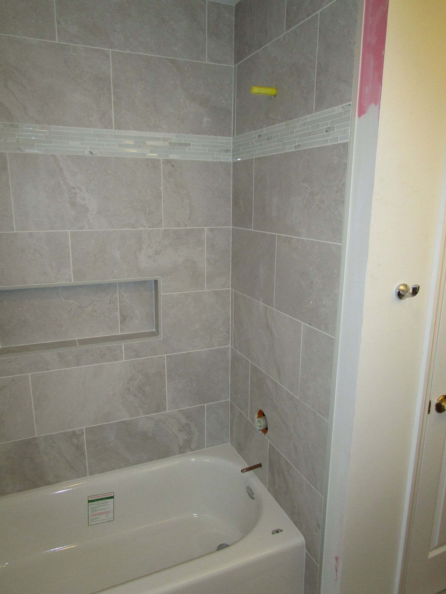 A bathroom shower under renovation with grey wall tiles, a built-in niche, and exposed plumbing fixtures above the tub.