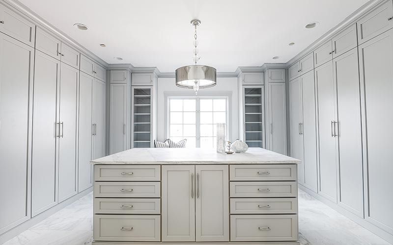 A spacious, light-gray walk-in closet featuring floor-to-ceiling cabinets, a central island with drawers, and a chandelier.