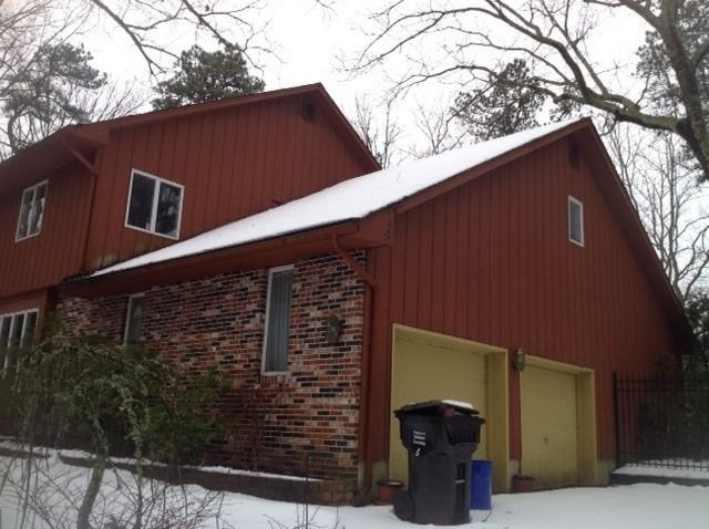 A two-story house with red vertical siding, a brick facade, and two garage doors, surrounded by snow in a winter setting.