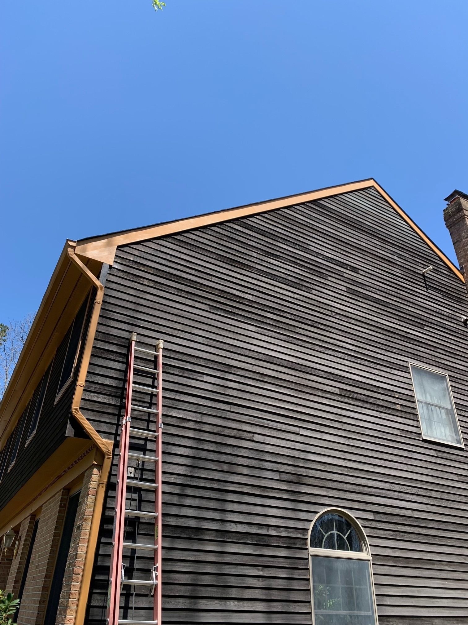 A tall ladder leans against the weathered, dark wood siding of a two-story home under a clear blue sky.