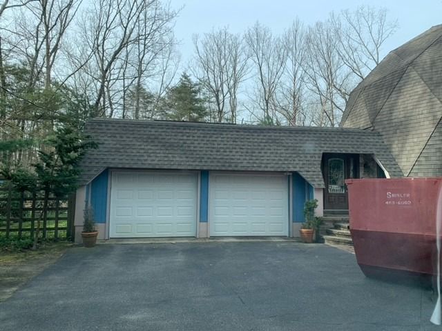 A two-car garage with white doors, blue trim, and a shingled roof, next to a large dome house and a red dumpster.