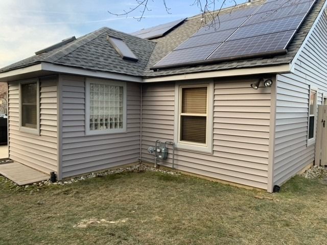 Light grey siding house with solar panels on a shingled roof, a skylight, and visible utility meters in a yard.