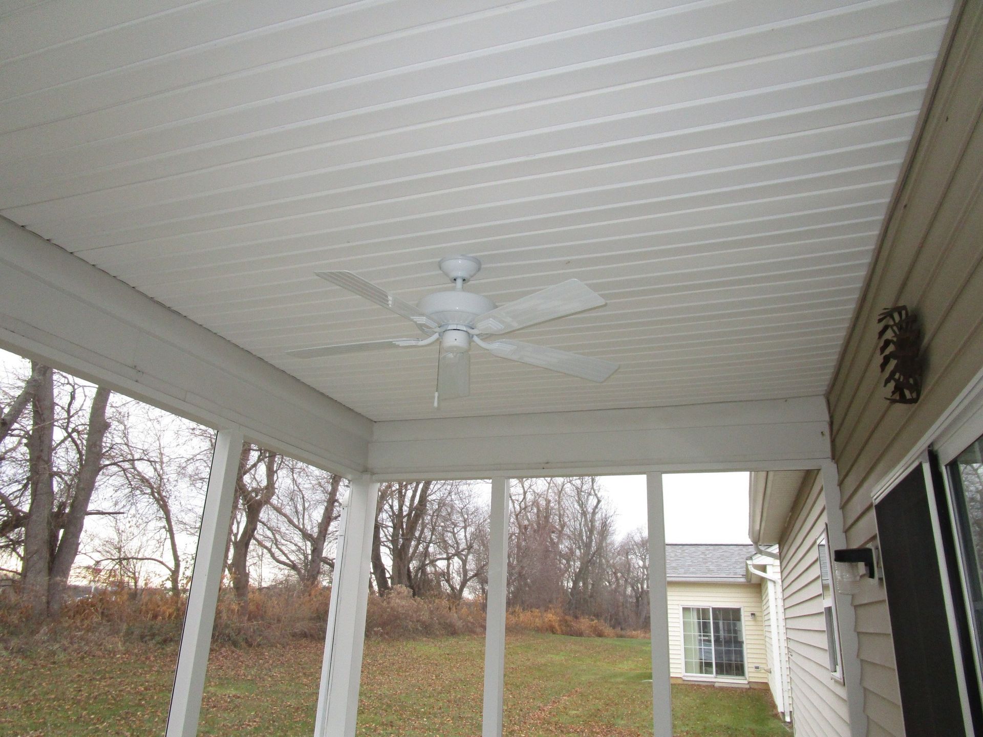 White ceiling fan mounted in a screened-in porch with a view of trees and another house in the distance.