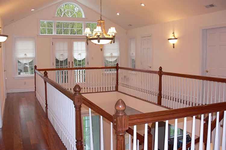 An interior view of a bright home balcony landing with wooden floors, white railings, and a chandelier under a vaulted roof.