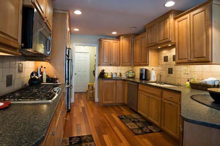 A bright kitchen featuring wooden cabinets, dark countertops, hardwood floors, and a view into an adjacent hallway.