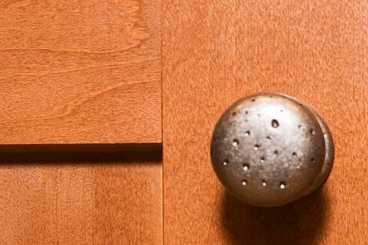 A round, pitted, metallic cabinet knob attached to a light-brown wood cabinet door.