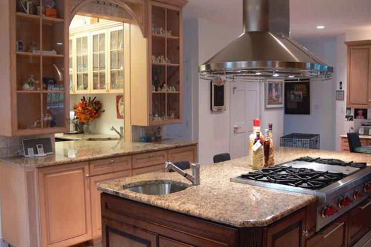 Kitchen island with a gas stovetop and sink in front of light wood cabinetry and glass-front upper cabinets.