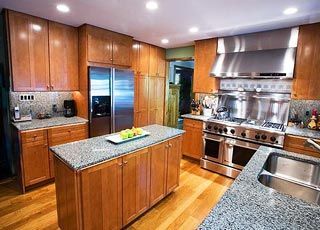A kitchen with wooden cabinets, stainless steel appliances, a granite-topped island, and light-colored hardwood floors.