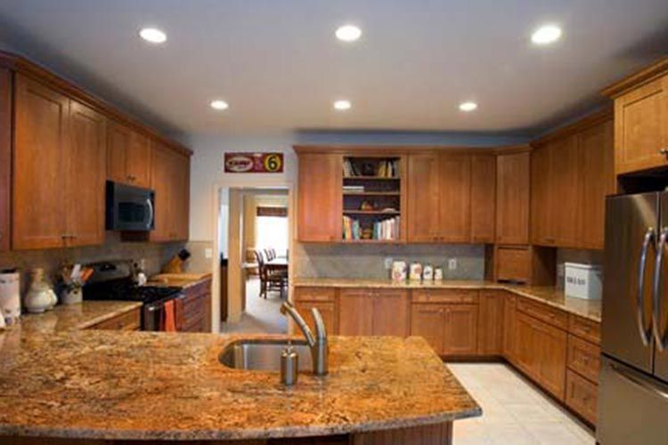 A well-lit kitchen featuring wood cabinets, granite countertops, a central island with a sink, and stainless appliances.