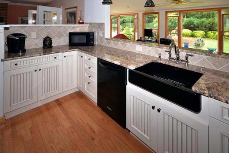 A bright kitchen with white beadboard cabinets, granite countertops, a black farmhouse sink, and hardwood flooring.