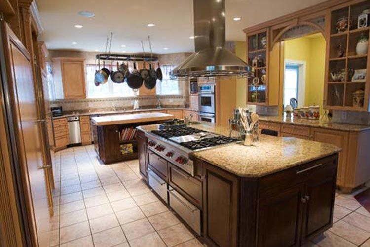A spacious kitchen featuring natural wood cabinets, granite countertops, a central island with a stove, and tiled floors.