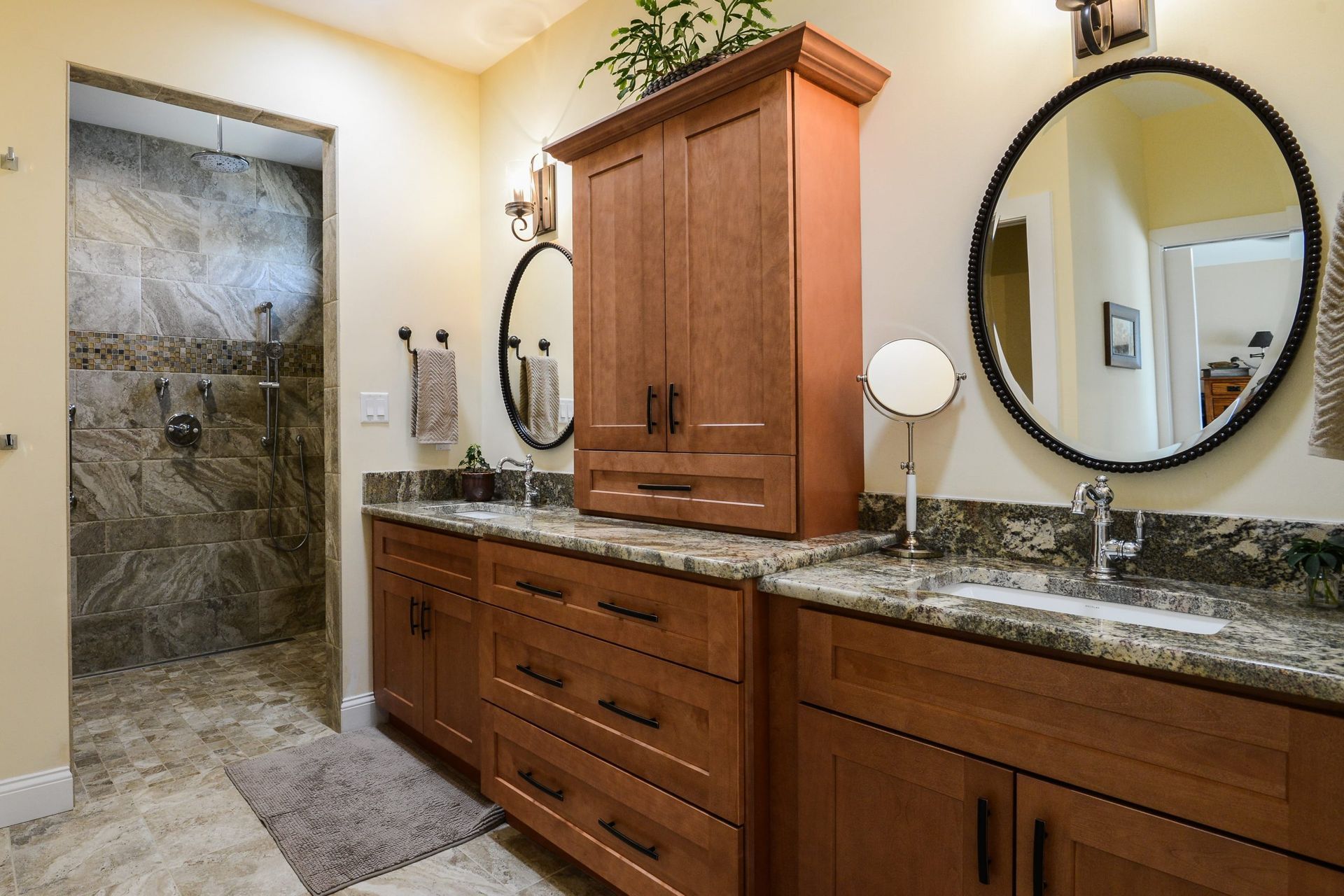 A master bathroom with wooden cabinetry, granite countertops, two round mirrors, and a tiled walk-in shower.