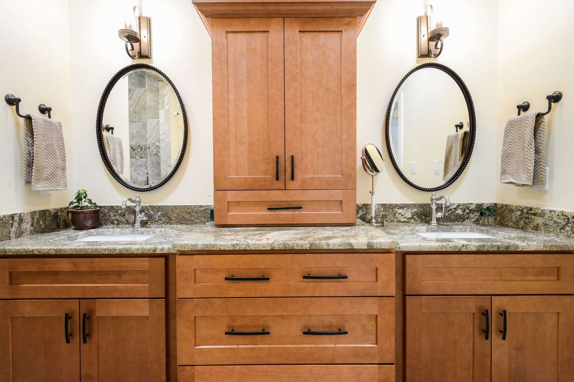 Double bathroom vanity with wood cabinets, granite countertops, dual oval mirrors, and wall-mounted light fixtures.