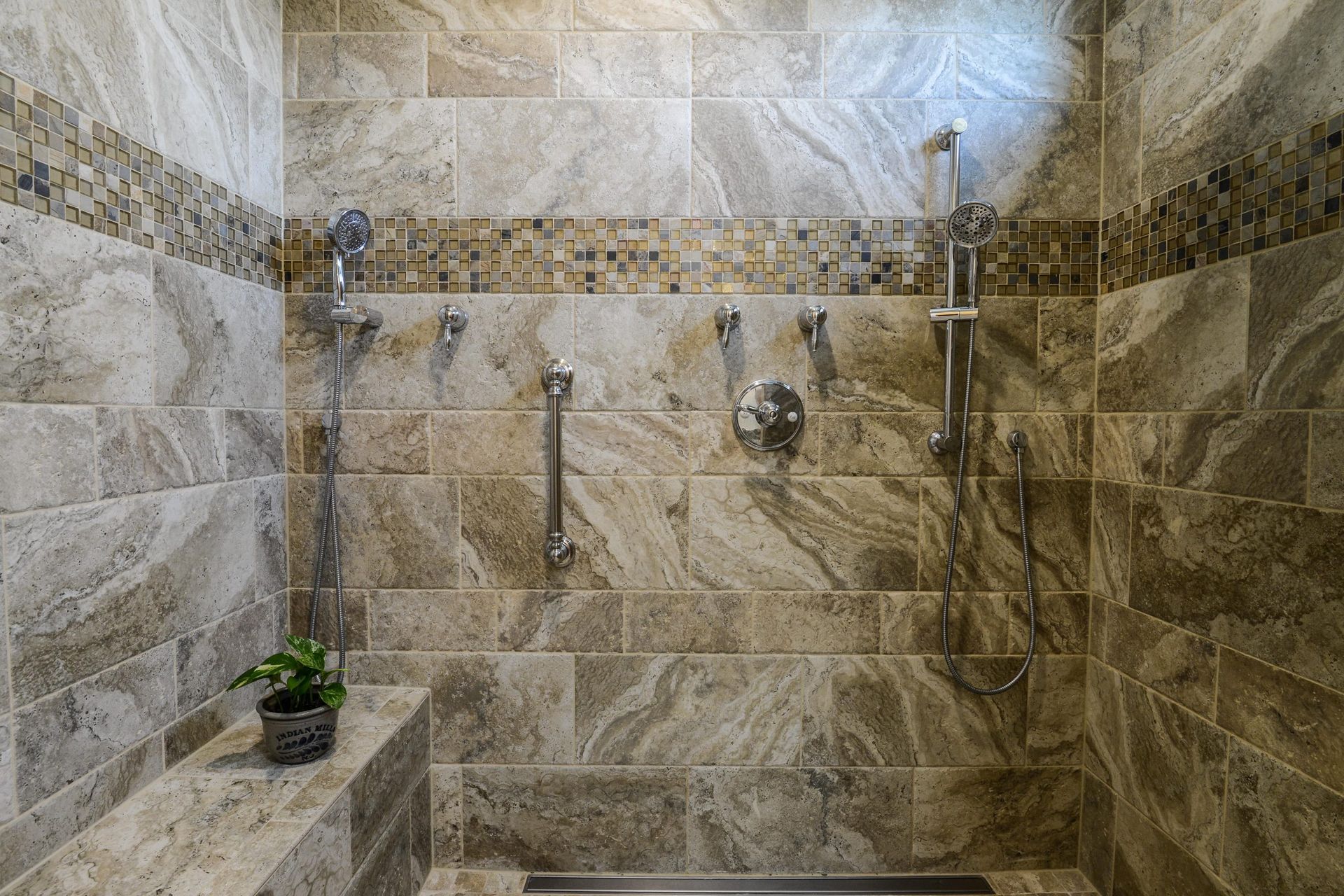 A stone-tiled walk-in shower with a built-in bench, a potted plant, and multiple chrome shower fixtures on the wall.