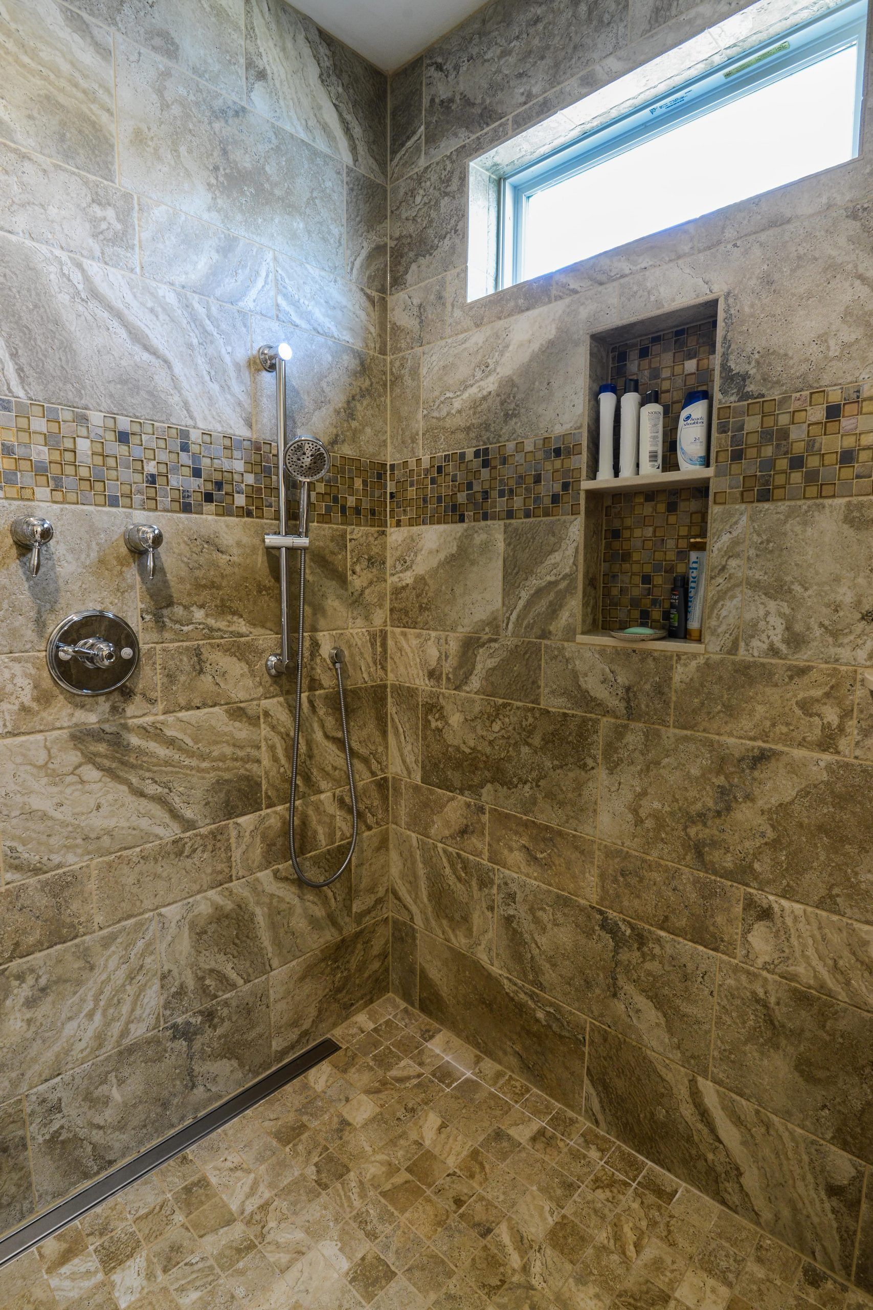 Modern shower with brown marble tile walls, a horizontal mosaic accent band, a built-in shelf, and a metal drain.