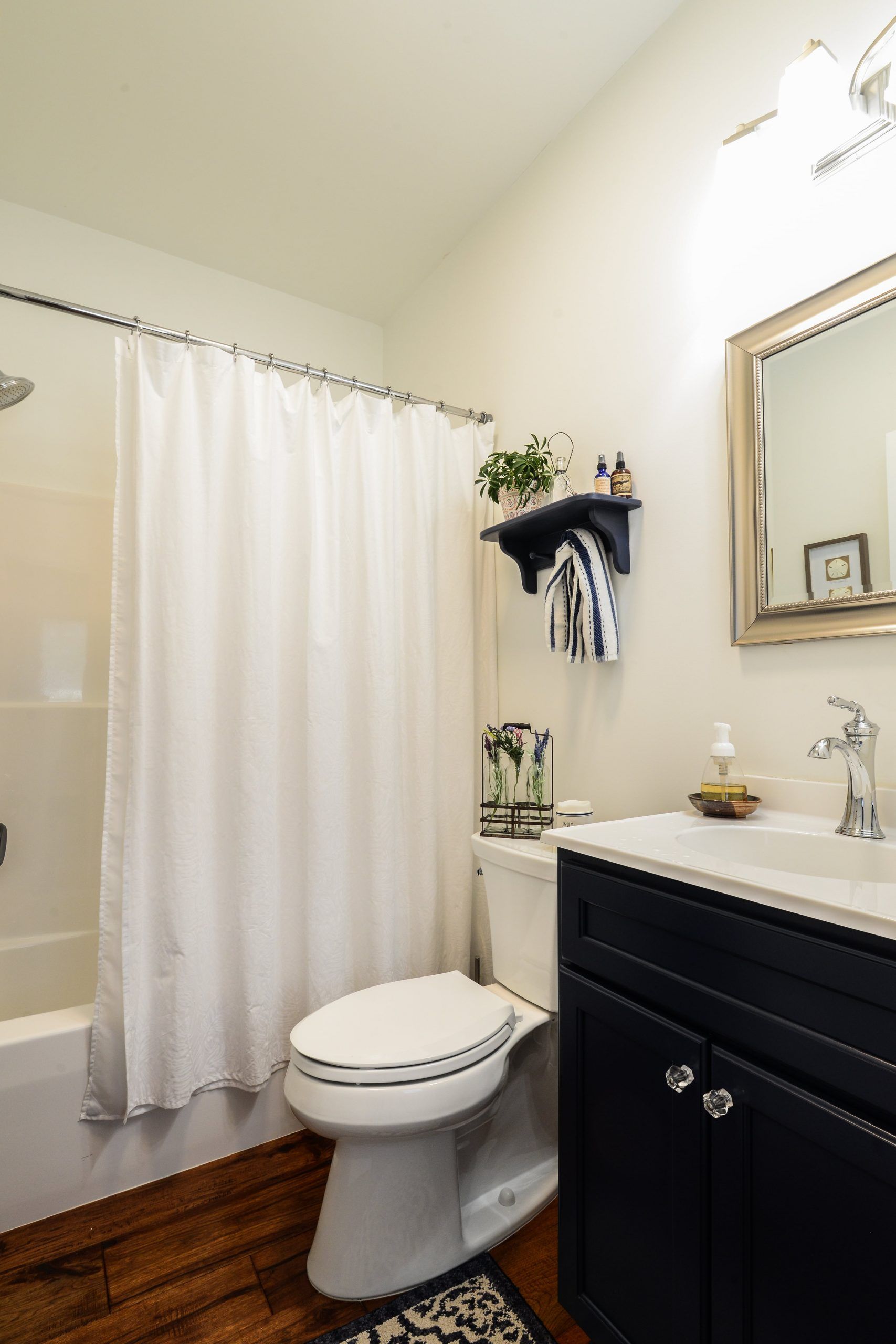 A modern bathroom with a dark blue vanity, a white toilet, a white shower curtain, and a wall-mounted shelf with decor.