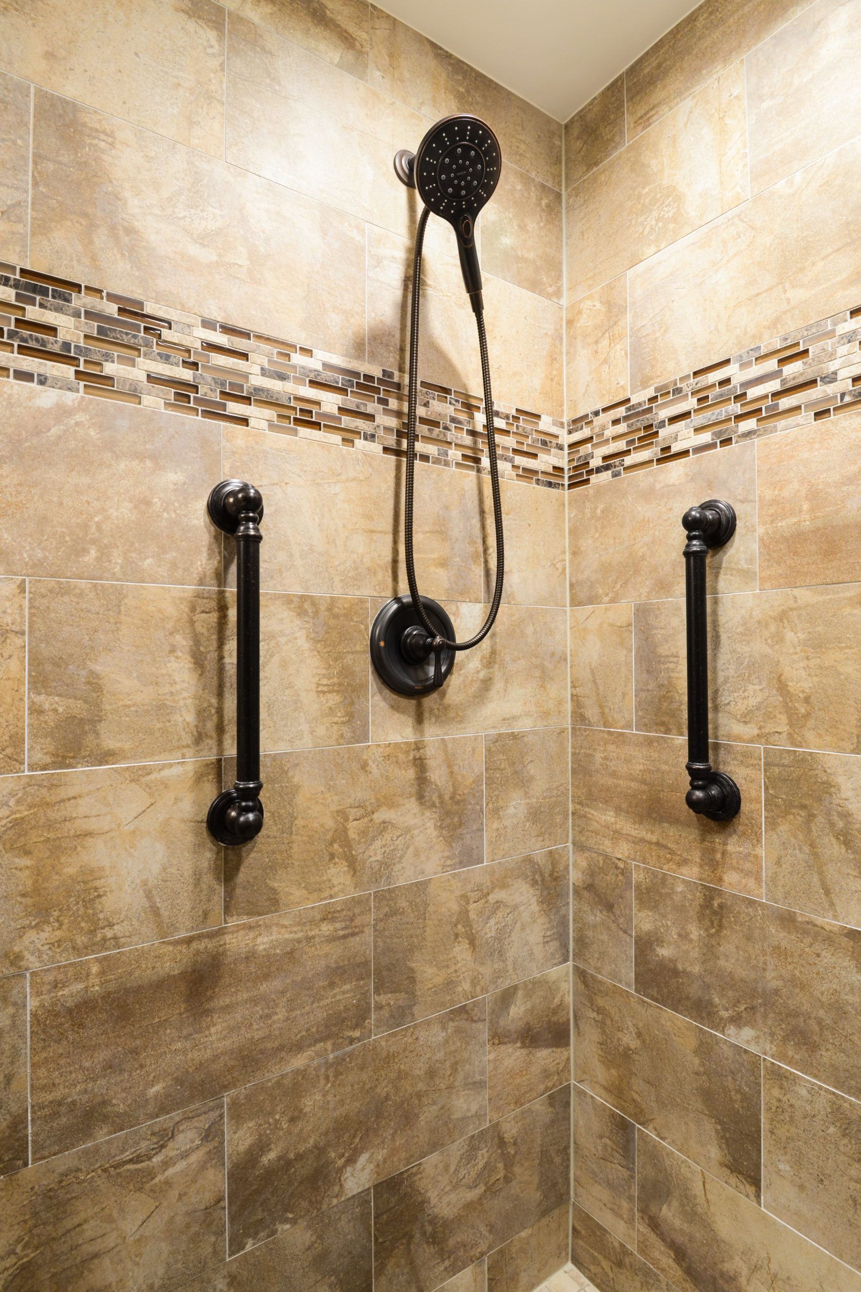A shower with two dark grab bars installed on tan tiled walls, flanking a central handheld showerhead.