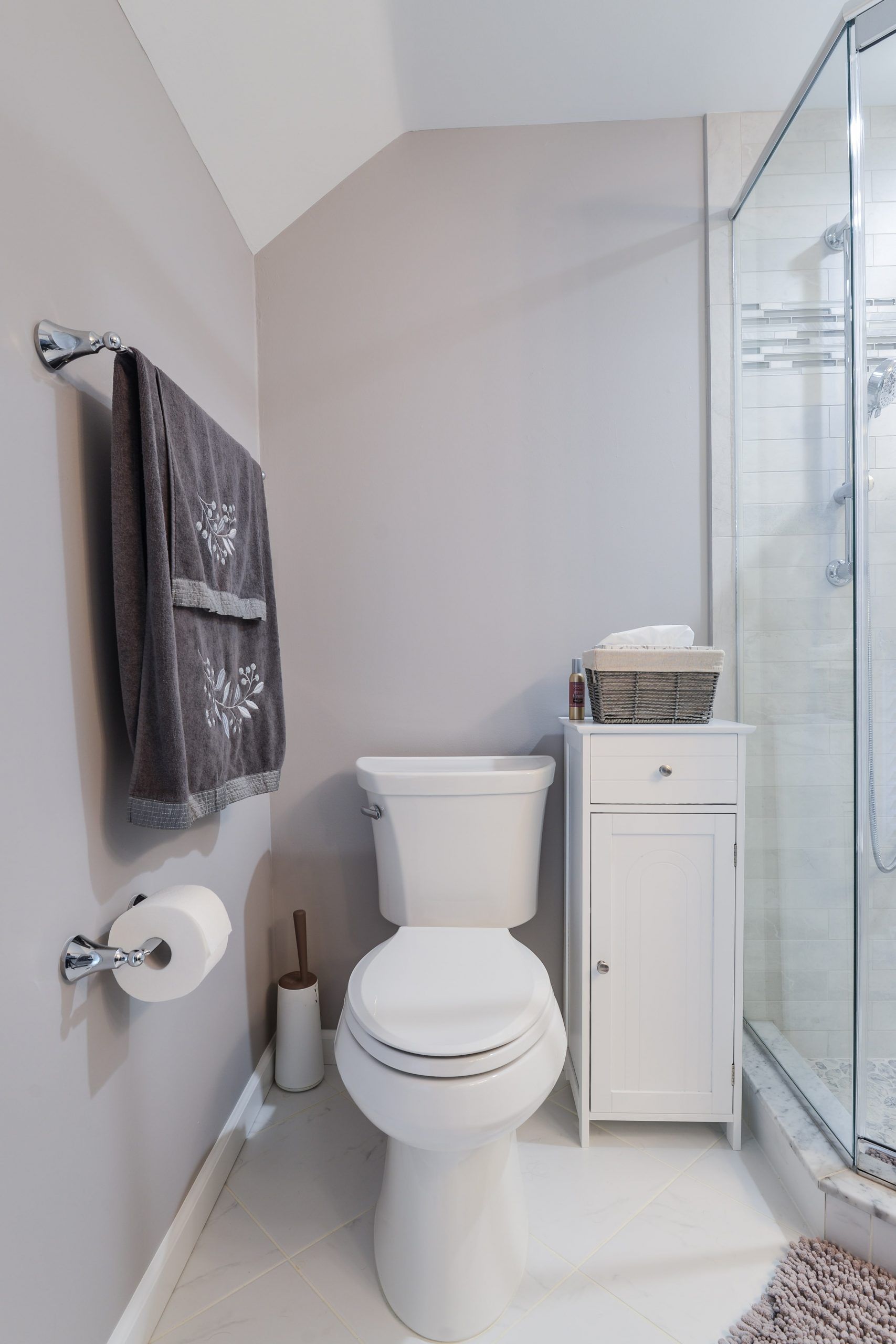 A modern bathroom featuring a white toilet, a tall white storage cabinet, a gray towel on the wall, and a glass shower.