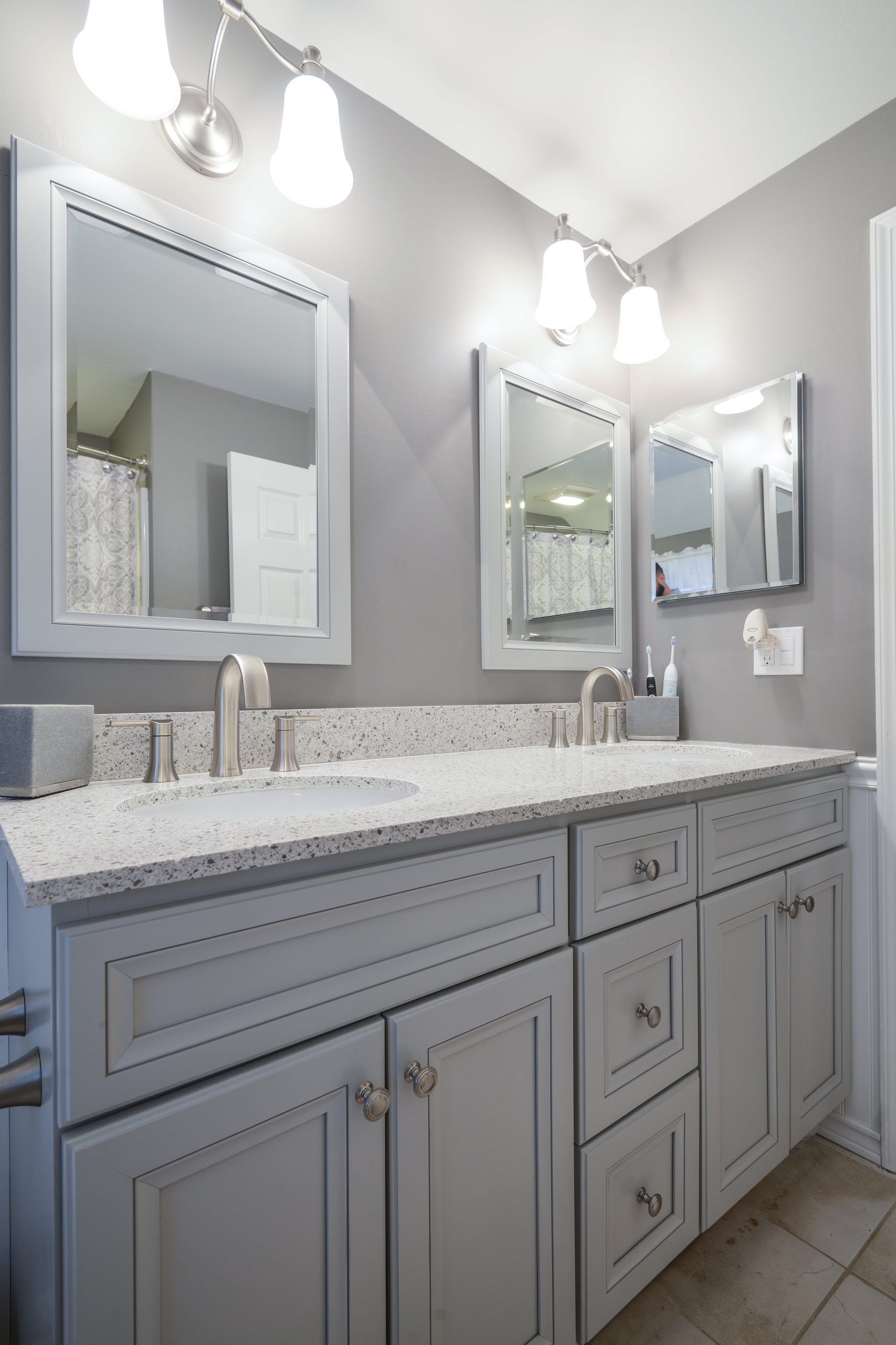 A bathroom vanity featuring grey cabinets, a speckled stone countertop with two sinks, and two framed mirrors above.