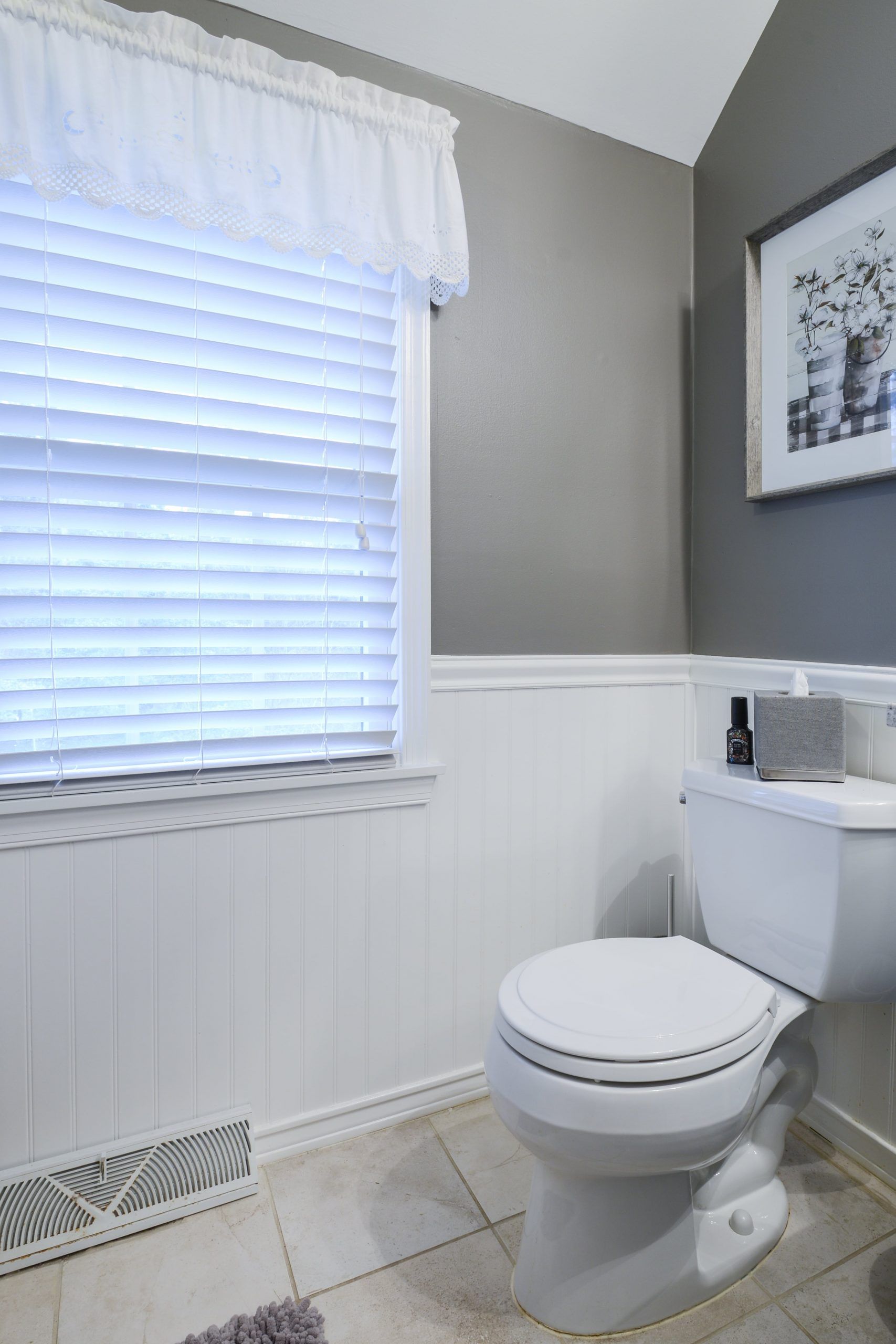 A white toilet sits in a small bathroom with gray walls above white wainscoting and a window with white blinds.