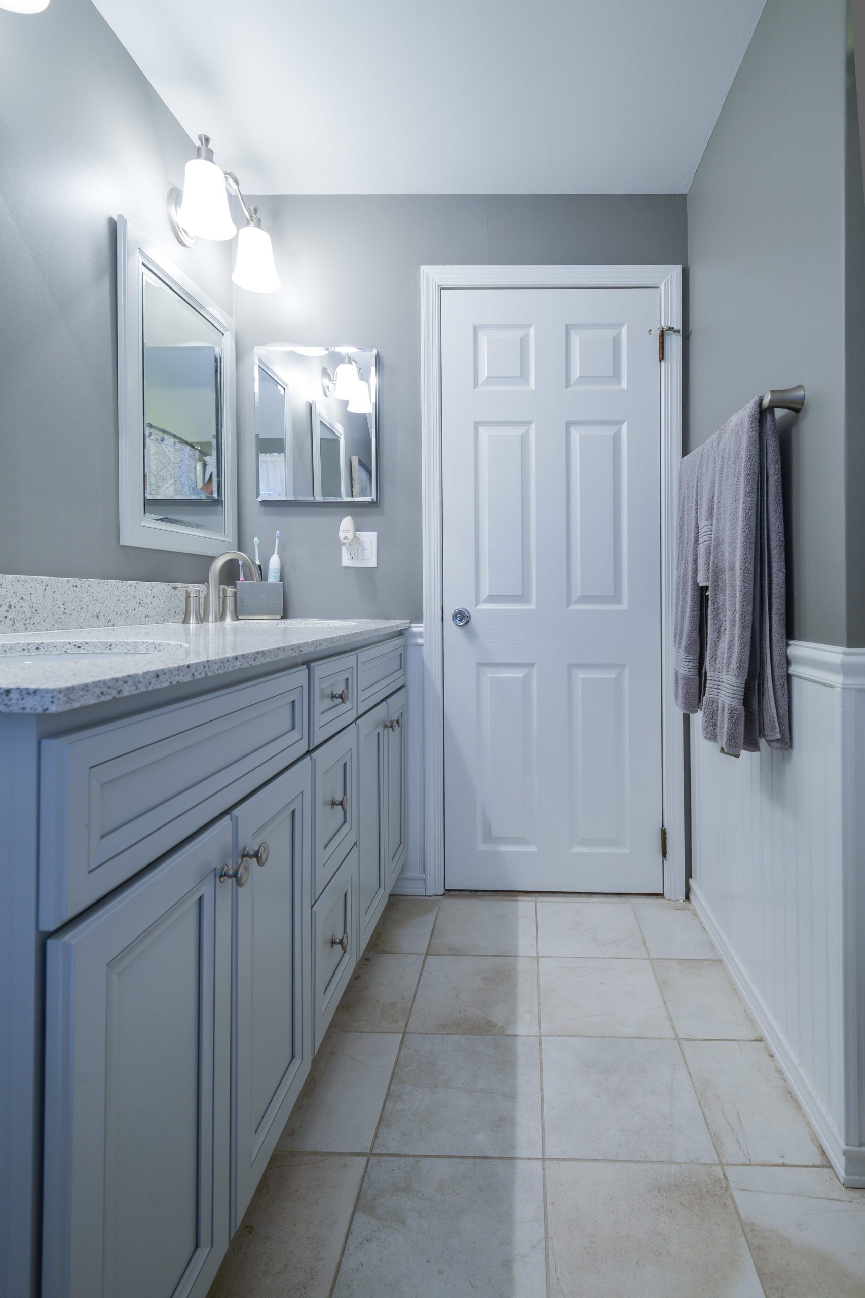 A modern bathroom with light gray walls, a white vanity with a granite countertop, a mirror, and a white interior door.